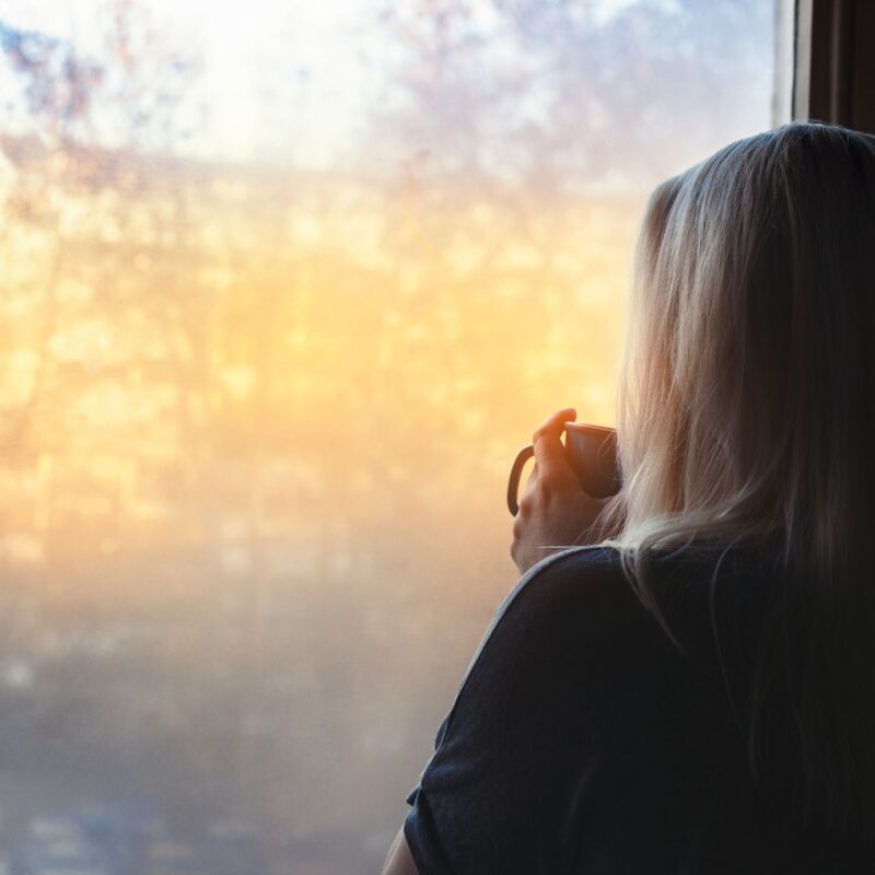 woman looking out of window with coffee mug cupped in both hands